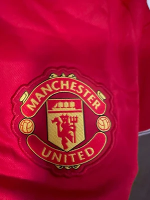 Manchester United matchtröja röd - Säljer en röd Manchester United matchtröja från Adidas med klubbmärke på bröstet, vita detaljer och sponsorlogga på framsidan. Tröjan har korta ärmar och klassisk krage med vita ränder på axlarna. Perfekt för fotbollsfans!