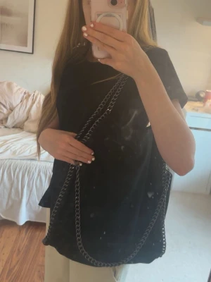 Svart axelväska med kedjedetaljer - Snygg svart axelväska med grova silverfärgade kedjor som axelrem och runt kanterna. Väskan har en mjuk, slouchy form och innerfack med dragkedja. Perfekt för dig som gillar en cool och trendig look.Ny pris 400 kr