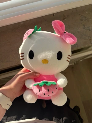 Hello Kitty mjukisdjur med jordgubbe - 25x15 cm Söt Hello Kitty mjukisdjur i vitt och rosa plysch. Katten har en rosa rosett och håller i en rosa jordgubbe med gröna blad. Perfekt som dekoration eller present till någon som gillar kawaii och samlarprylar.