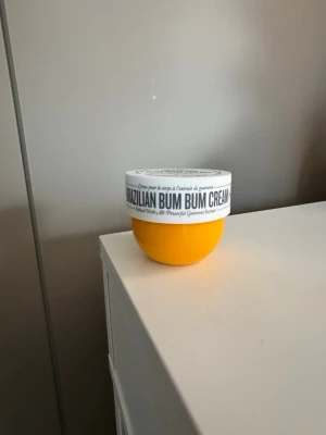 Sol de Janeiro  Bum Bum Cream 62 - En ikonisk Brazilian Bum Bum Cream från Sol de Janeiro i en rund, gul burk med vitt lock. Krämen är känd för sin snabbabsorberande formula och doft av pistage och salt karamell. Perfekt för att återfukta och ge huden glow.