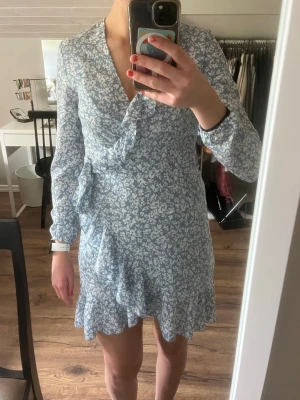 Blå blommig omlottklänning - Söt blå klänning med vitt blommönster och volangdetaljer. Klänningen har omlottmodell, v-ringning och långa ärmar.