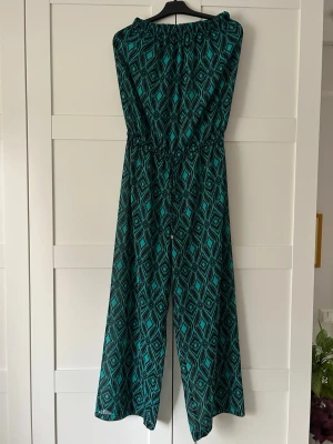 Offshoulder jumpsuit - Snygg jumpsuit med grönt och svart grafiskt mönster. Modellen har vida ben, knytband i midjan och är offshoulder. Den är lite genomskinlig och ett perfekt plagg att snabbt dra över på stranden efter att man tagit ett dopp.