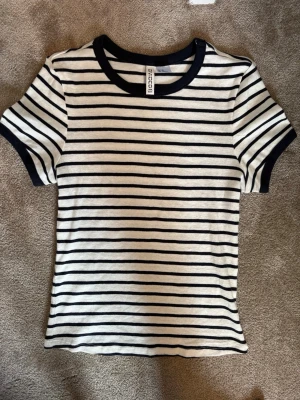 Randig t-shirt från H&M  - Säljer en vit och marinblå randig t-shirt från Divided. T-shirten har korta ärmar, rund halsringning med marinblå kant och är gjord i mjuk bomull. Perfekt till jeans eller kjol för en enkel och stilren look.
