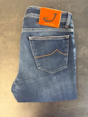 Jacob Cohën Style 688, stl 33 - Snygga blå slim fit jeans med orange sömmar och en unik orange patch.  Jeansen har klassisk femficksdesign och lätt slitna partier framtill för en trendig look. Perfekta för dig som gillar en modern och avslappnad stil.