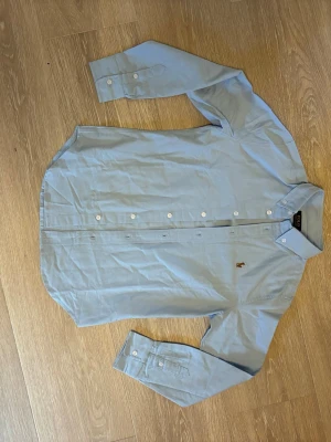 Ljusblå skjorta från Polo Ralph Lauren - Klassisk ljusblå skjorta från Polo Ralph Lauren med broderad logga på bröstet. Skjortan har knappar framtill, button-down krage och långa ärmar. Perfekt för en clean och stilren look.