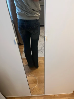 Mörkblå bootcut jeans med brodyr - Säljer ett par mörkblå bootcut jeans med snygg brodyr på bakfickorna. Jeansen har en klassisk femficksmodell och lätt slitna detaljer för en cool look. Tillverkade i stretchigt denimtyg som sitter skönt och formar sig efter kroppen.
