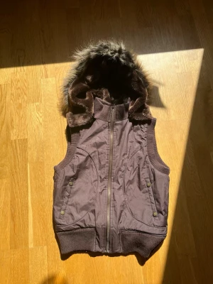 Brun väst med pälsluva  - Snygg brun väst med dragkedja framtill, ribbade muddar och två fickor med tryckknappar. Storlek 8 och passar som S 