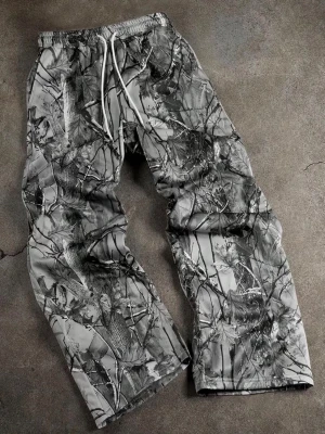 Grå camo mjukisbyxor med snörning - Säljer ett par grå mjukisbyxor med camouflagemönster och vita dragsnören i midjan. Byxorna har avslappnad passform, elastisk midja och raka ben. Perfekta för chill dagar eller streetwear-stil. Ny och storlek M
