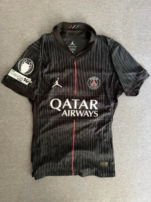PSG Player Version - Säljer en svart PSG matchtröja med diskreta ränder och röd detalj framtill. Tröjan har Qatar Airways-tryck, PSG-logga och Jordan-logga på bröstet samt Champions League-märke på ärmen. Tillverkad i Nike’s Dri-Fit ADV material, perfekt för fotboll eller träning.