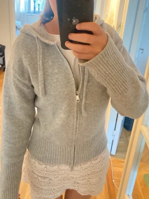Ljusgrå Zip hoodie från Design by Amor - Mysig ljusgrå hoodie från Design by Amor med dragkedja framtill och ribbade muddar. Tröjan har huva med dragsko och är gjord i ett mjukt stickat material som känns riktigt skönt mot huden. Perfekt för dig som gillar en clean och enkel stil.