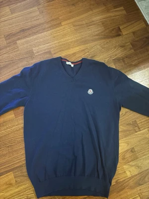 Gammal model Mörkblå Moncler V-ringad tröja - Gammal model Snygg mörkblå V-ringad tröja från Moncler med diskret logotyp på bröstet. Tröjan har långa ärmar och ribbade muddar vid ärmslut och nederkant. Klassisk och stilren design med röd detalj på insidan av kragen.