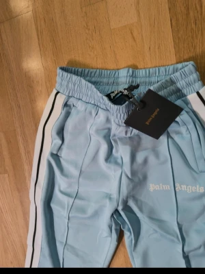 Palm Angels ljusblå byxor - Snygga ljusblå byxor från Palm Angels med vita och svarta ränder längs sidorna. Byxorna har elastisk midja, dragkedja vid benslut och logga tryckt på ena benet. Perfekta för en chill och trendig look. Stl är M men passar i L med 