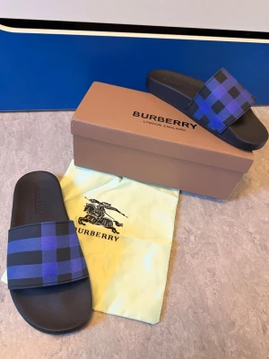 Blå-rutiga tofflor från Burberry - Snygga tofflor från Burberry med blå och svarta rutor på remmen. Tillverkade i syntetmaterial med klassisk slip-on design. Perfekta för chill dagar hemma eller på semestern. Kommer med originalkartong och dustbag.