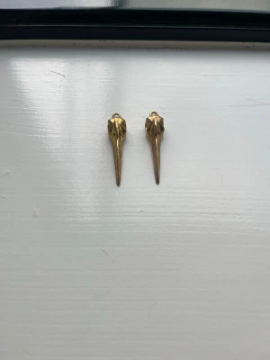 Pilgrim guldiga berlocker i fågelskalleform - Unika örhängen i form av fågelskallar i guldfärgad metall. De har en cool och edgy design med lång näbb och detaljerade former. Perfekta för dig som vill sticka ut med din stil.