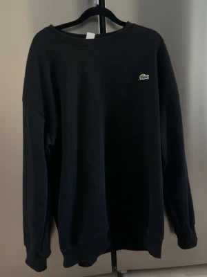 Svart sweatshirt från Lacoste - En stilren svart sweatshirt från Lacoste med rund halsringning och diskret krokodillogga på bröstet. Tröjan har långa ärmar och ribbade muddar vid ärmslut och nederkant. Perfekt för en avslappnad och trendig look.