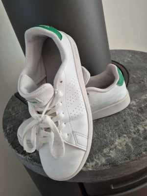 Vita Adidas sneakers med gröna detaljer - Klassiska vita Adidas sneakers med gröna detaljer på hälen och logga. Skorna har lågt skaft, vita snören och perforerade ränder på sidorna. Tillverkade i syntetmaterial med gummisula. Perfekta för en clean och sportig stil. Skorna är I bra använd skick inga hål och så...