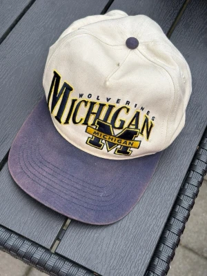 Michigan Wolverines keps beige/blå - Snygg keps i beige med blått och gult Michigan Wolverines-brodyr framtill. Skärmen är blå och kepsen har en klassisk form med justerbar baksida. Perfekt för dig som gillar college-stil och sportiga accessoarer.