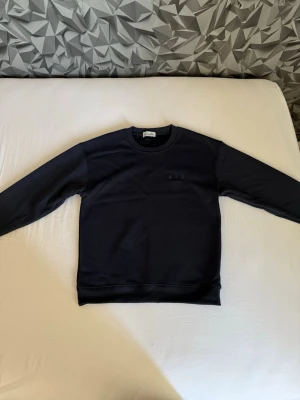 Mörkblå sweatshirt med rund hals - En stilren mörkblå sweatshirt med rund halsringning och långa ärmar. Tröjan har en diskret broderad logga på bröstet och ribbade muddar vid ärmslut och nederkant. Perfekt för en avslappnad och trendig look.