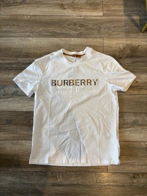 Vit burberry t-shirt med logga - Snygg vit t-shirt från burberry med klassisk logga i guld och svart text på bröstet. Modellen har rund halsringning och korta ärmar. Perfekt för dig som gillar stilrena och exklusiva plagg med tydlig branding.