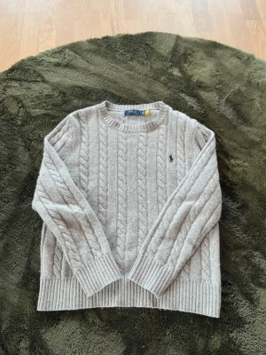 Grå kabelstickad tröja Polo Ralph Lauren - Kabelstickad grå tröja från Polo Ralph Lauren med rund halsringning och broderad marinblå logga på bröstet. Tröjan har ribbstickade muddar och nederkant, perfekt för lager-på-lager och snygg till jeans.