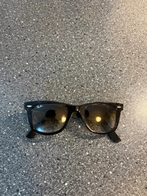 Ray-Ban Solglasögon - Klassiska svarta Wayfarer solglasögon från Ray-Ban med bred båge och mörka glas. Snygg och tidlös design som passar till det mesta. Ray-Ban-loggan syns tydligt på ena linsen. Perfekta för soliga dagar.