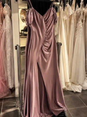 Festklänning  - Elegant långklänning i lila satin med smala axelband och draperad framsida. Klänningen har en djup slits framtill och en vacker glansig yta som ger en lyxig känsla. Perfekt för speciella tillfällen.