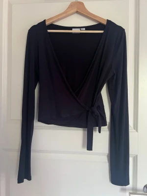 Marinblå omlottopp från Gina Tricot - Marinblå omlottopp från Gina Tricot med långa ärmar och knyt i sidan. Toppen har en djup v-ringning och är tillverkad i ett mjukt, stretchigt material som sitter snyggt på kroppen. Perfekt att styla med jeans eller kjol.