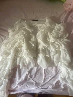 Vit volangkofta med långa ärmar - Supersöt vit kofta med massor av volanger och långa ärmar. Koftan har en luftig och flowy känsla, perfekt för att lyfta vilken outfit som helst. Materialet är tunt och lätt, vilket ger en snygg och trendig look.
