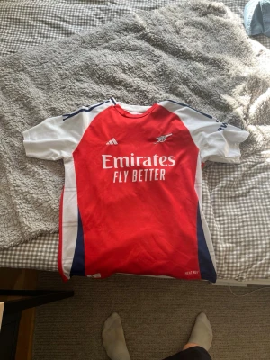 Arsenal matchtröja Ødegaard #8 - Snygg Arsenal matchtröja i rött, vitt och marinblått med Ødegaard och nummer 8 på ryggen. Tröjan har Adidas-logga, klubbmärke och Emirates Fly Better-tryck på bröstet. Tillverkad i lätt och ventilerande material, perfekt för fotboll eller träning.