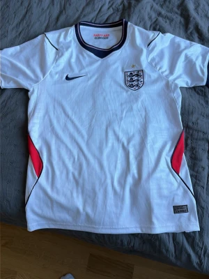 Englands vm tröja - Säljer en vit England fotbollströja från Nike med broderat landslagsemblem och en guldstjärna på bröstet. Tröjan har marinblå detaljer vid kragen och röda inslag på sidorna. Perfekt för dig som älskar fotboll och vill stötta England.