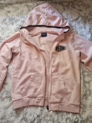 Ljusrosa zip hoodie från PeakPerformance - Ljusrosa hoodie med dragkedja framtill och två fickor. Har en huva med svart kant och ett tryck på bröstet. Tillverkad i mjukt sweatshirtmaterial, perfekt för chill dagar. Storlek 160.