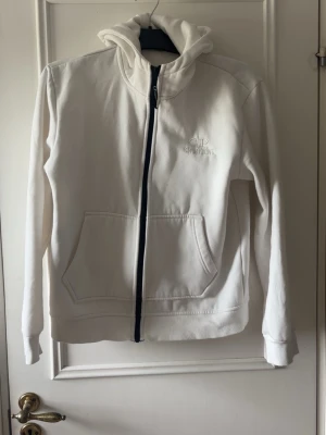 Cp company kofta - Säljer en vit hoodie med dragkedja och huva som har inbyggda glasögon. Hoodien har fickor framtill och broderad logga på bröstet. Perfekt för dig som gillar streetwear och vill sticka ut med en unik detalj på huvan.