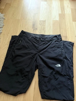 Svarta Frilufts byxor från The North Face - Säljer ett par svarta friluftsbyxor för dam i strl 6 (S) från The North Face. Byxorna har rak passform, bälteshällor och sidofickor. Tillverkade i ett slitstarkt och lätt material som passar perfekt för utomhusaktiviteter. Loggan är tryckt på vänster lår.