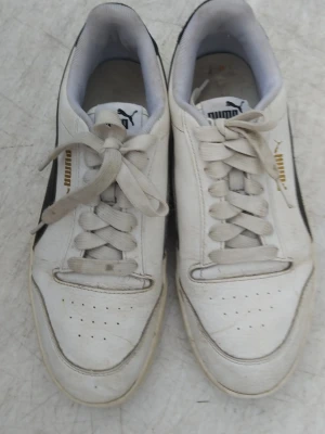 Vita Puma sneakers med svart detalj - Snygga vita sneakers från Puma med klassisk svart sidodetalj och guldfärgad logga. Skorna har lågt skaft, vita snören och perforerade detaljer på tån. Ovandelen är i läder och insidan är fodrad i grått tyg. Perfekta för en sportig och avslappnad stil. Pris kan diskuteras 