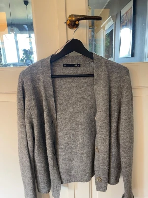 Grå stickad kofta från 157 - Mysig grå stickad kofta från 157 med v-ringad öppning och knappar framtill. Perfekt att slänga över en t-shirt eller topp för en chill och enkel look. Koftan har lång ärm och en klassisk passform.