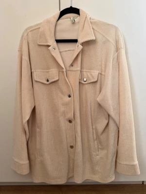 Beige overshirt från NLY Trend - Stilren beige overshirt från NLY Trend i mjuk manchester. Skjortjackan har klassisk krage, tryckknappar framtill, två bröstfickor med lock och sidofickor. Perfekt lager på lager-plagg med avslappnad passform och uppvikta ärmslut.