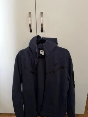 Mörkblå Nike hoodie med dragkedja - Säljer en mörkblå hoodie från Nike med dragkedja framtill och svart Nike-logga på bröstet. Hoodien har en huva och diskreta svarta detaljer över bröstet. Perfekt för en avslappnad och sportig stil.