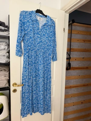 Blå blommig långklänning - Blå långklänning med vita blommor och trekvartsärm. Klänningen har krage och knappar framtill. Tillverkad i mjuk viskos som ger ett skönt fall. Perfekt för dig som gillar en feminin och avslappnad stil.