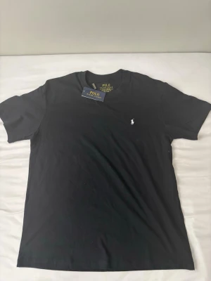 Svart t-shirt från Polo Ralph Lauren - Klassisk svart t-shirt från Polo Ralph Lauren med den ikoniska broderade loggan på bröstet. T-shirten har rund hals och korta ärmar, tillverkad i mjuk bomull för en skön känsla. Perfekt basplagg för alla tillfällen.