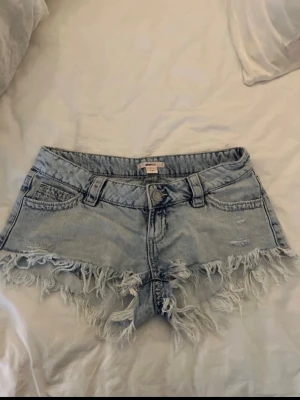 Ljusblå lowwaist jeansshorts - Säljer ett par nya ljusblå  jeansshorts med fransig kant och slitna detaljer. Shortsen har fem fickor, bälteshällor och knappstängning framtill. Perfekta för sommaren och ger en avslappnad vibe.