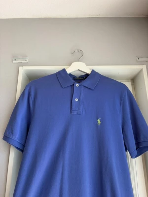 Lila  pikétröja från Ralph Lauren - Klassisk lila pikétröja från Ralph Lauren med korta ärmar och krage. Tröjan har två knappar framtill och den ikoniska broderade loggan i gult på bröstet. Perfekt till jeans eller shorts för en avslappnad men snygg stil.