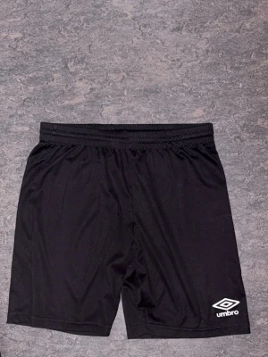 Svarta träningsshorts från Umbro - Svarta träningsshorts från Umbro med elastisk midja och logga på ena benet. Tillverkade i ett lätt och svalt polyestermaterial som passar perfekt för sport och aktivitet. Klassisk och enkel design som är lätt att matcha.