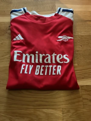 Arsenal röd matchtröja Adidas - Säljer en röd Arsenal matchtröja från Adidas med vita och blå detaljer. Tröjan har trycket 'Emirates Fly Better' på bröstet och Arsenal-loggan på vänster sida. Tillverkad i ett lätt och ventilerande material, perfekt för träning eller match.