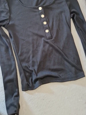 Mörkgrå ribbad långärmad topp - Snygg mörkgrå ribbad topp med lång ärm och rund halsringning. Toppen har fyra silvriga knappar framtill som en snygg detalj. Perfekt att styla med jeans eller kjol för en chill look.