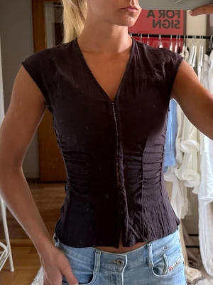 Brun v-ringad blus med knappar - Snygg brun blus med v-ringning och knappar framtill. Modellen är figurnära med korta ärmar och lätt rynkade sömmar i midjan för en smickrande siluett. 