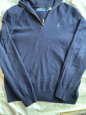 Mörkblå stickad tröja Polo Ralph Lauren - Mörkblå stickad tröja från Polo Ralph Lauren med halv dragkedja och broderad logga på bröstet. Tröjan har ribbade muddar och hög krage, perfekt för lager på lager. Klassisk och stilren design i mjukt material.