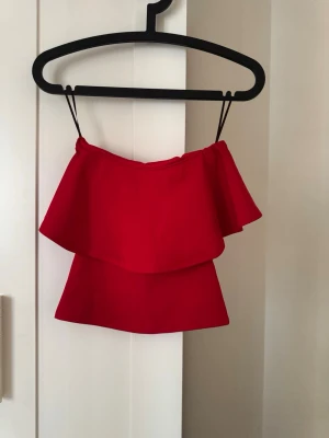 Röd volangig axelbandslös topp H&M - Snygg röd axelbandslös topp från H&M med volangdetalj. Perfekt för dig som vill sticka ut med en färgstark och trendig look. Toppen har en croppad passform och tunna axelband. Superfin till både jeans och kjol!