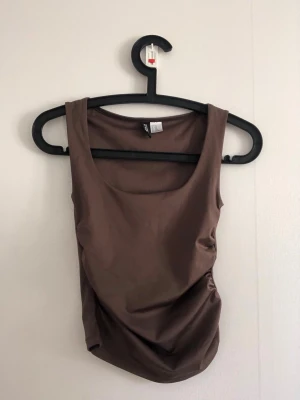 Brun ärmlös linnetopp - Snygg brun ärmlös linnetopp med rund halsringning och figurnära passform. Perfekt att styla med jeans eller kjol för en enkel och trendig look. Tillverkad i mjukt material som känns skönt mot huden.