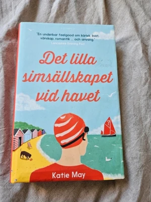 Det lilla simsällskapet vid havet - En charmig feelgood-roman av Katie May om vänskap, kärlek och att våga börja om. Omslaget är färgglatt med illustration av strand, badhytter och en person i röd badmössa. Perfekt för dig som gillar lättsam och varm skönlitteratur.