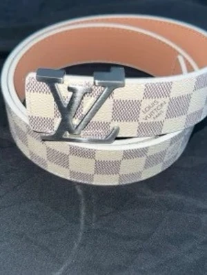 Rutig IV skärp i vitt - Snyggt bälte från Iv i klassiskt damiermönster i beige och vitt. Bältet har ett stort IVspänne i silverfärgad metall och insidan är ljusbrun. Perfekt accessoar för att lyfta vilken outfit som helst.